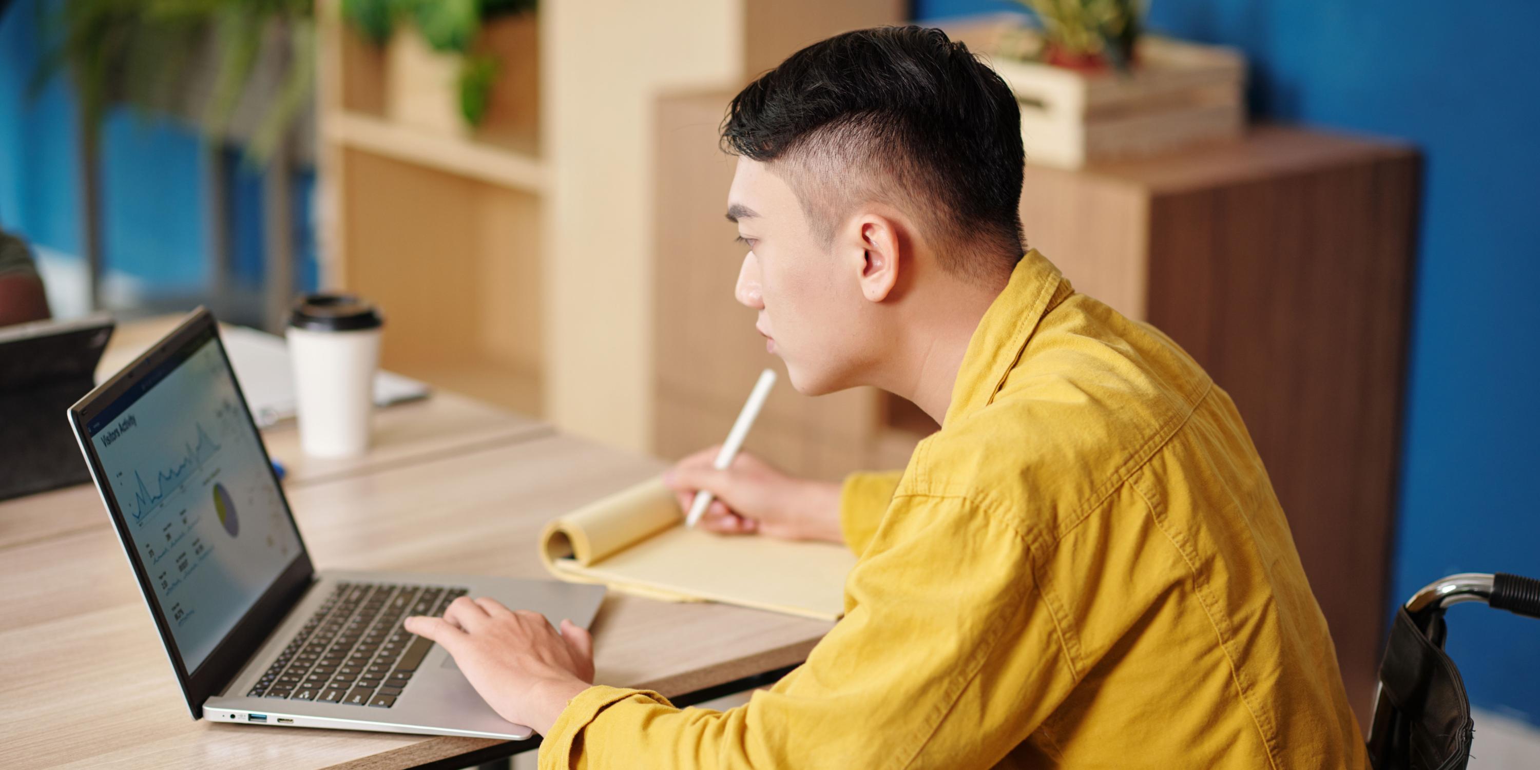 A person wearing a yellow shirt looking at their laptop screen which is showing a slide with various graphs. He is also holding a pen with his right hand to take notes with on his notebook.
